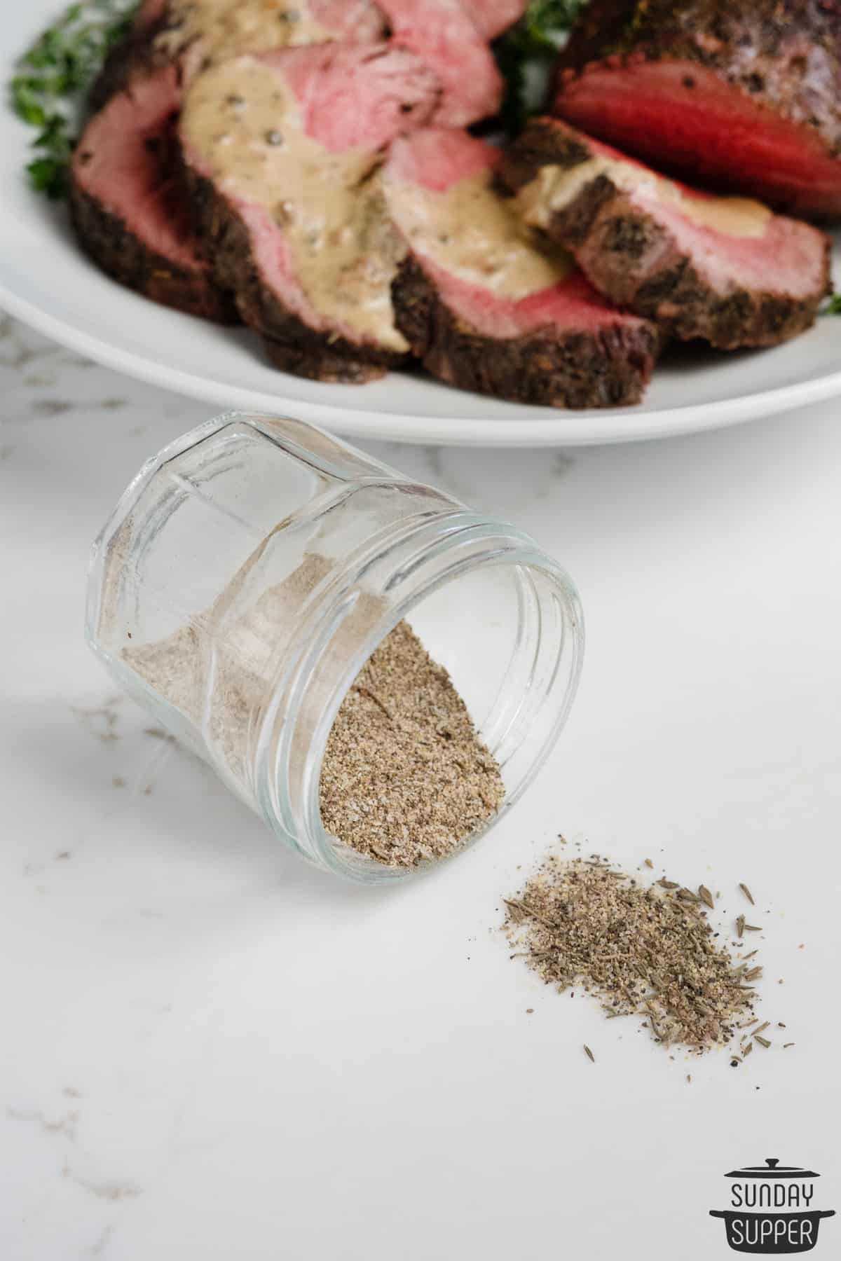 beef tenderloin rub spilling out of a glass jar next to cooked beef tenderloin on a plate