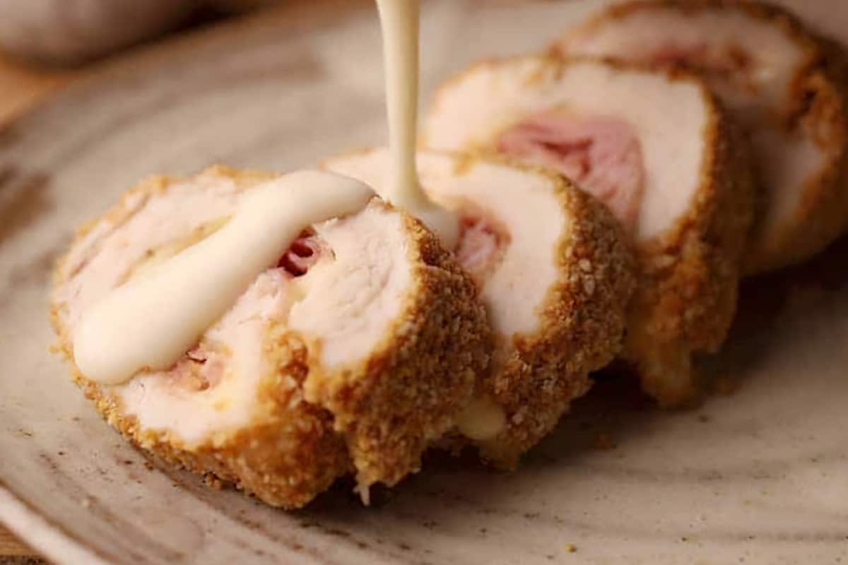 pouring parmesan sauce over chicken cordon bleu sliced on a plate