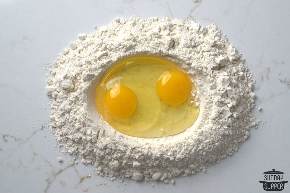 a mound of flour on a clean working surface with two eggs cracked into a well in the middle