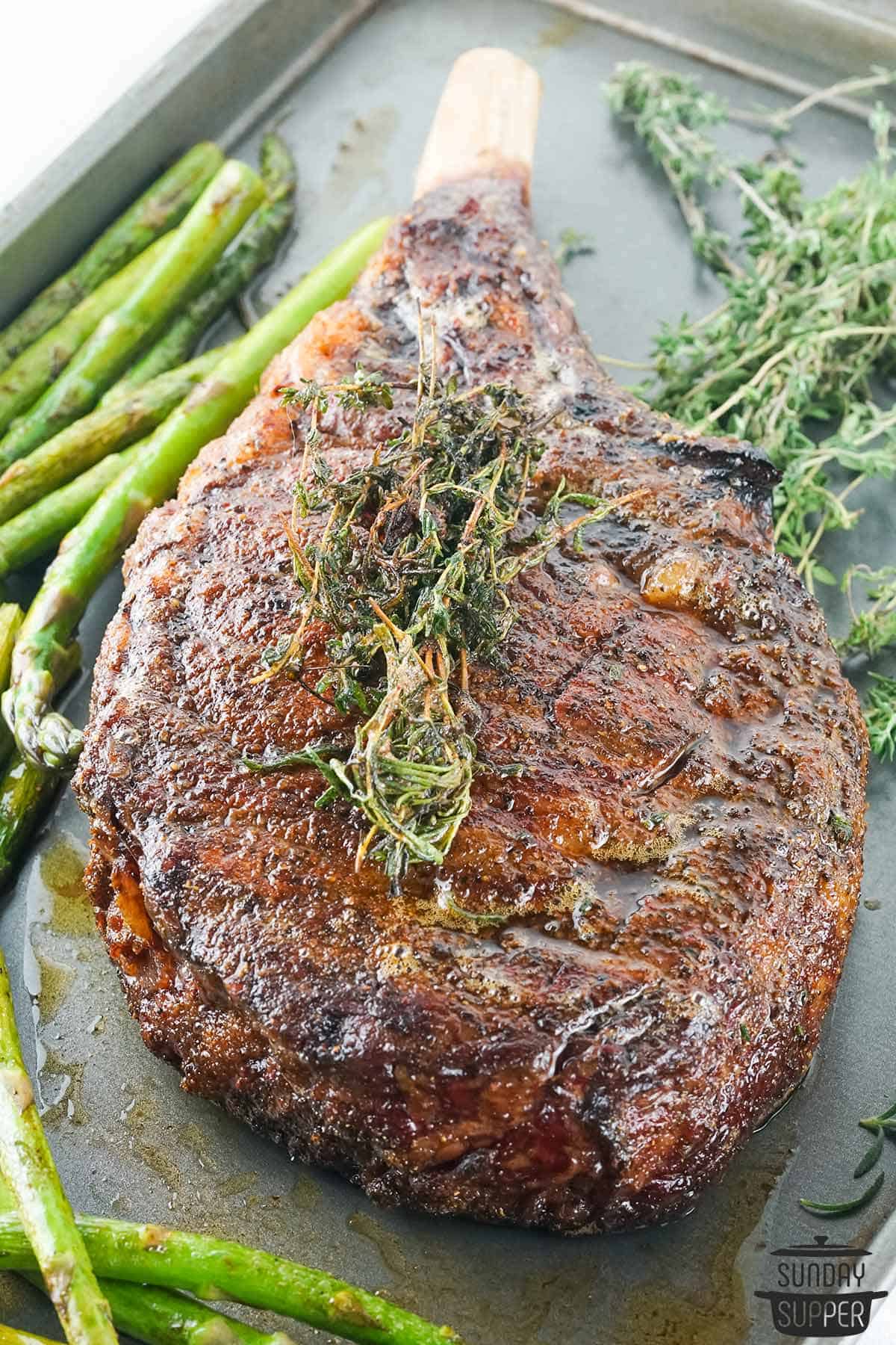smoked tomahawk steak on a baking sheet with asparagus and thyme