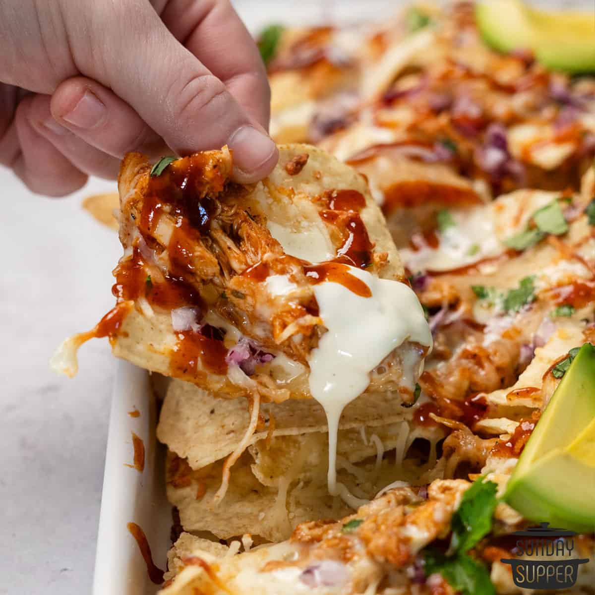 a hand taking a chip loaded with bbq chicken and queso off a tray of nachos