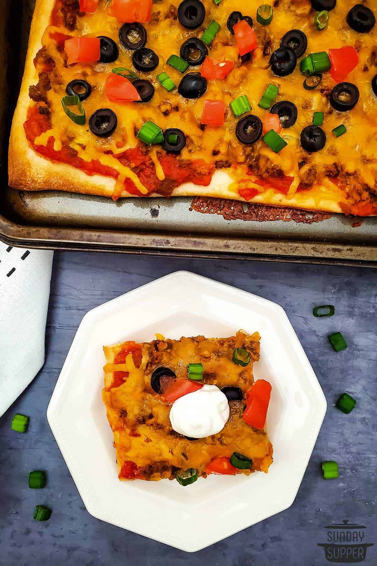 a slice of taco pizza on a plate next to a pan of pizza