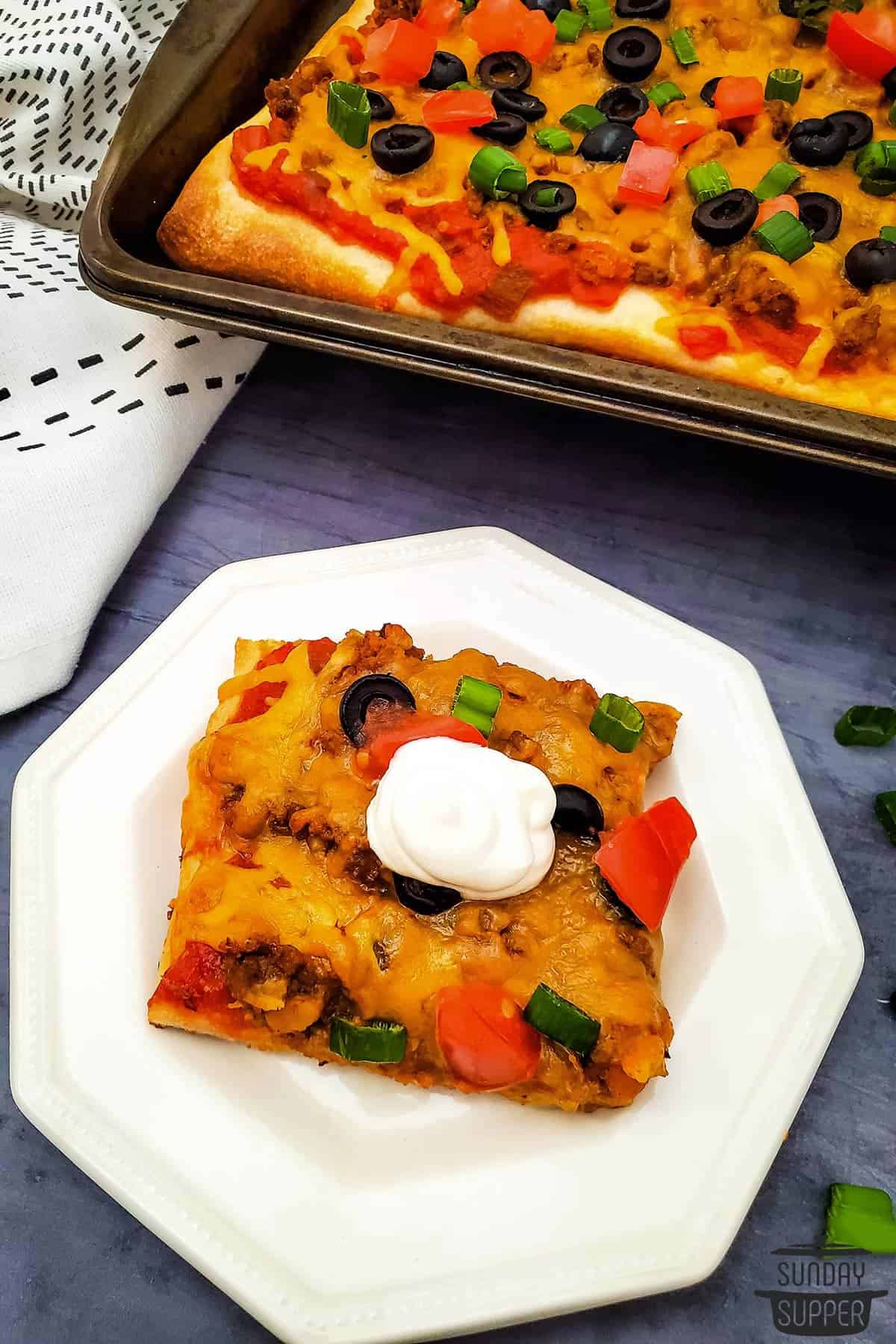 a slice of taco pizza on a plate next to the pan of pizza