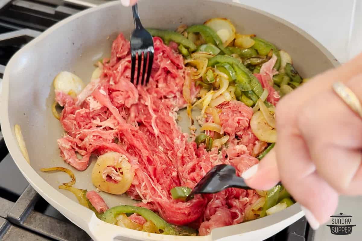 separating shredded ribeye with two forks in a pan